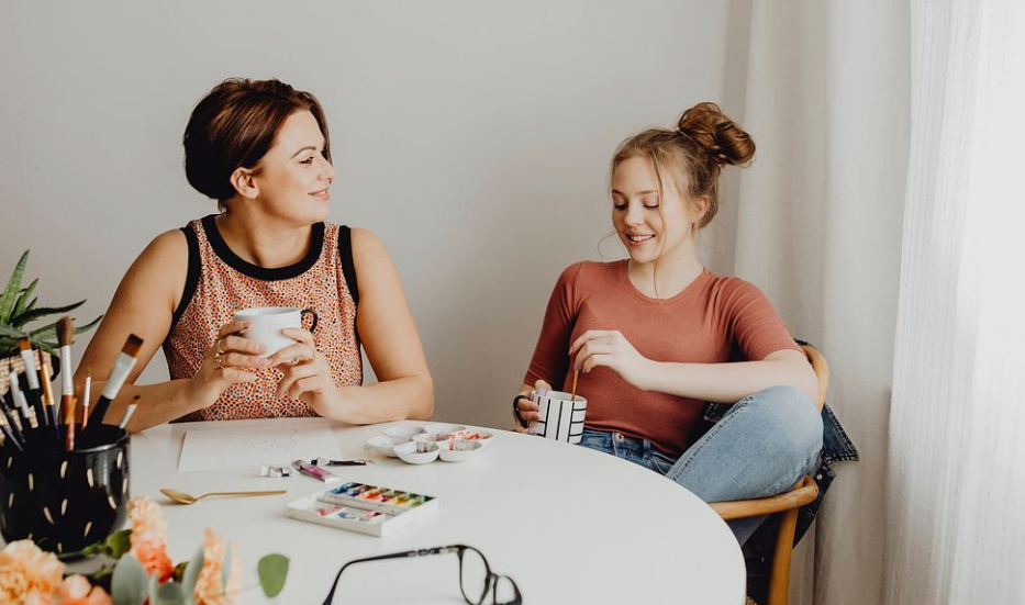 "Two women sitting at a table, smiling and having a casual conversation while holding cups. Art supplies, including brushes and paints, are spread out on the table, suggesting a creative or relaxing activity."