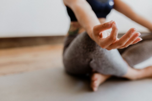 "Person sitting in a cross-legged yoga pose with one hand in a meditation gesture, focusing on relaxation and mindfulness in a calm indoor space."