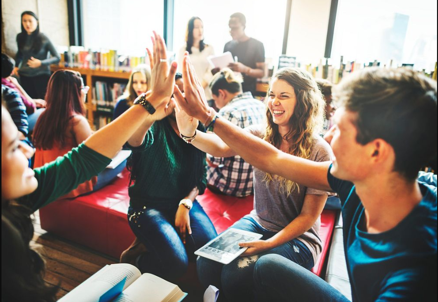 A group of young unverisity students from diverese backgrounds sitting in a classroom. They are sitting in a circle of chairs close together. They're smiling and high fiveing each other